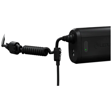 A close-up view of the black LED Lenser headlamp's battery pack and its connecting cable. The coiled cable is attached to the battery pack, which has a green LED indicator showing the charge level. The background is white, highlighting the details and design of the battery pack and its connection points.