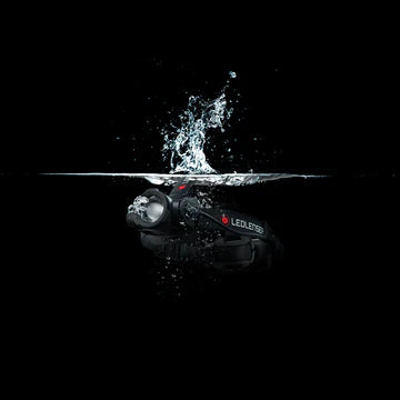 A black LED Lenser headlamp partially submerged in water, with a splash effect captured at the water's surface. The image emphasizes the headlamp's water-resistant or waterproof capabilities. The LED Lenser logo is visible on the headlamp's strap. The background is completely black, highlighting the splash and the headlamp.