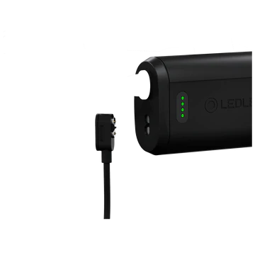 A close-up view of a black LED Lenser battery pack being charged. The charging cable with a connector is positioned near the battery pack, ready to be plugged in. The battery pack has green LED indicators showing the charge level. The "LED LENSER" branding is visible on the side of the battery pack.