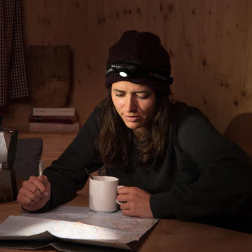 A woman wearing a black LED Lenser headlamp and a dark beanie is sitting at a wooden table indoors. She is reading a map, with a white mug and a pen in her hands. The headlamp is turned on, illuminating the map and her work area. The background features wooden walls, books, and other items, indicating a cozy indoor setting.