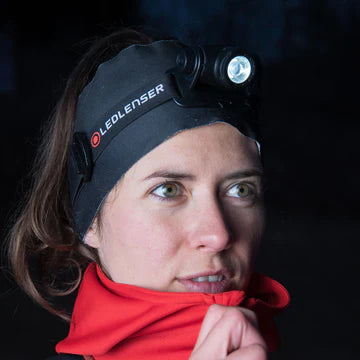 A close-up of a woman wearing a LED Lenser headlamp and a black headband with the LED Lenser logo. She is outdoors in a dark environment, wearing a red jacket and looking to the side. The headlamp is turned on, illuminating her face and the area in front of her.