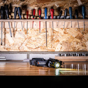 A black LED Lenser headlamp is placed on a wooden workbench in a workshop. The headlamp is turned on, emitting a beam of light. The background features a wall with various tools hanging, including screwdrivers, files, and other hand tools. The workbench has a neat and organized appearance, emphasizing the headlamp's utility in a workshop setting.