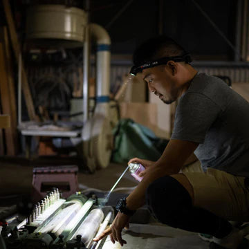 A man wearing a LED Lenser headlamp is kneeling in a workshop, inspecting or repairing a piece of machinery. The headlamp illuminates his work area, allowing him to see clearly in the dimly lit environment. The workshop is filled with various tools and equipment, indicating a setting for detailed technical or mechanical work.