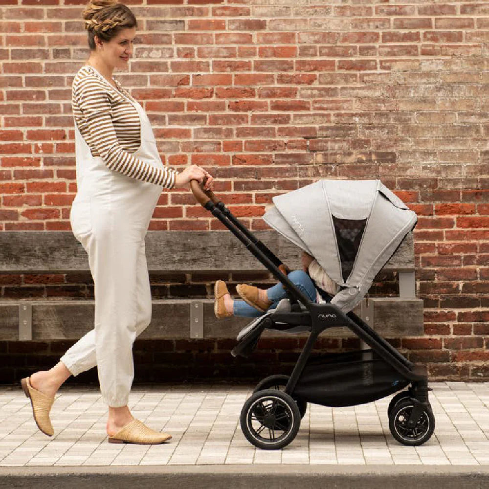 A woman pushes a Nuna stroller with a toddler seated inside, featuring a grey canopy and large wheels, while walking along a paved sidewalk in front of a brick wall.