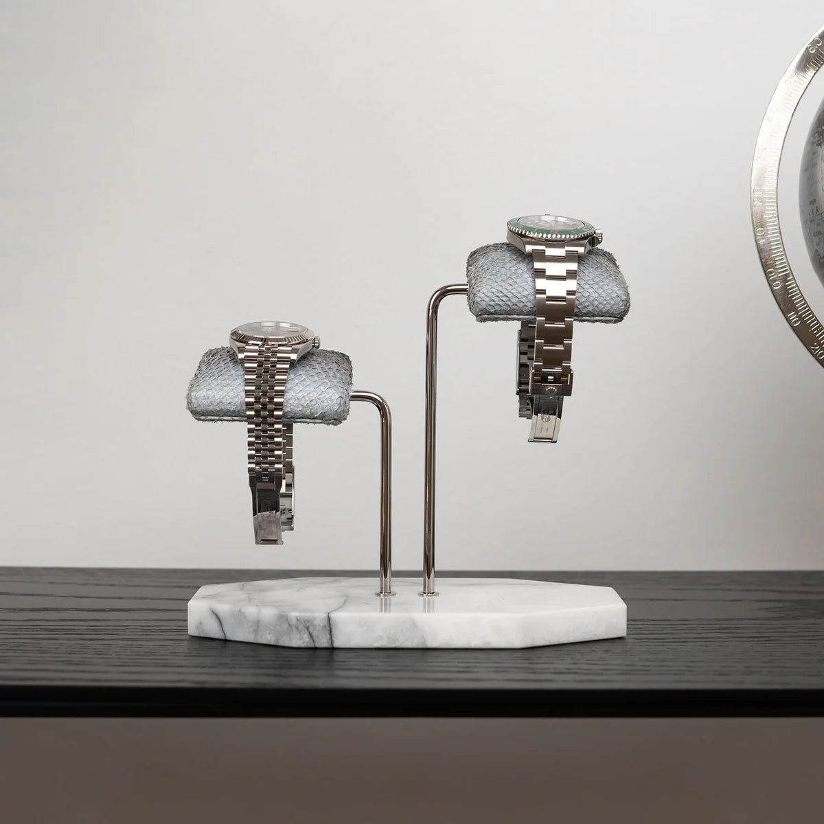 Two luxurious wristwatches are displayed on gray textured cushions, mounted on a double-arm stand with a white marble base. The stand features polished silver supports, adding to the elegant design. One watch has a silver metal band with a silver dial, while the other has a silver metal band with a green bezel. The display is set on a dark wooden surface against a plain white wall, creating a sophisticated and stylish presentation.