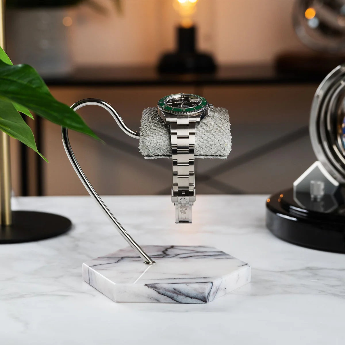 A wristwatch with a silver metal band and a green bezel is elegantly displayed on a textured gray cushion. The cushion is mounted on a sleek, curved metal stand with a hexagonal white marble base that features gray veining. The display is set on a white marble surface, and the background includes modern decor elements, such as a plant, a gold lamp, and another watch display, creating a sophisticated and stylish ambiance.