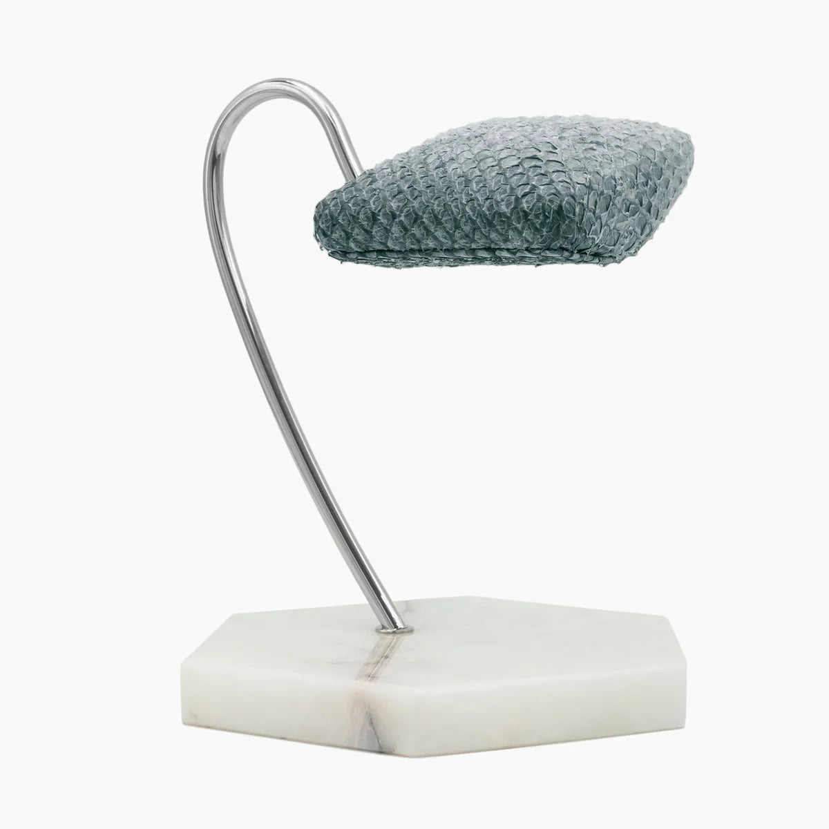 A display stand featuring a gray textured cushion mounted on a sleek, curved metal support. The base of the stand is a hexagonal white marble slab with subtle gray veining. The design is minimalist and elegant, ideal for showcasing a wristwatch or similar item. The contrast between the textured cushion, shiny metal support, and marble base creates a sophisticated and modern aesthetic.