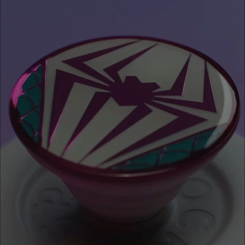 A top-down view of a PopSocket phone grip featuring a Spider-Man inspired design. The design consists of a stylized white spider emblem on a pink background with blue accents on the sides. The circular surface showcases the vibrant colors and striking emblem prominently.