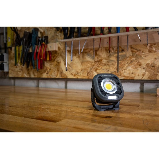 A SOLIDline LED work light placed on a wooden workbench in a workshop setting. The workbench has various tools hanging on a pegboard in the background, including pliers, screwdrivers, and other hand tools. The LED light is turned off, showcasing its compact and sturdy design.