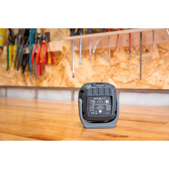 The back view of a SOLIDline LED work light placed on a wooden workbench in a workshop setting. The back panel displays various specifications, certifications, and a charging port. The workbench has various tools hanging on a pegboard in the background, including pliers, screwdrivers, and other hand tools.