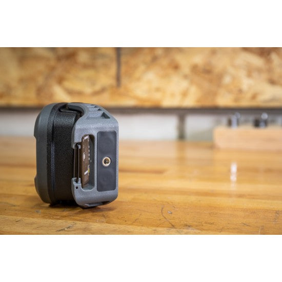 A close-up side view of a SOLIDline LED work light placed on a wooden workbench in a workshop setting. The image focuses on the adjustable stand and mounting hole of the work light. The background includes a blurred view of tools hanging on a pegboard.
