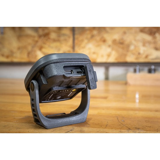 A close-up back view of a SOLIDline LED work light placed on a wooden workbench in a workshop setting. The image focuses on the charging port and USB ports, with the protective cover open. The background includes a blurred view of tools hanging on a pegboard.