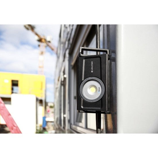 A LEDLenser portable light attached to the exterior wall of a building, emitting a bright beam. The background shows a construction site with a crane, buildings, and blue sky, highlighting the light's utility in outdoor and construction settings.