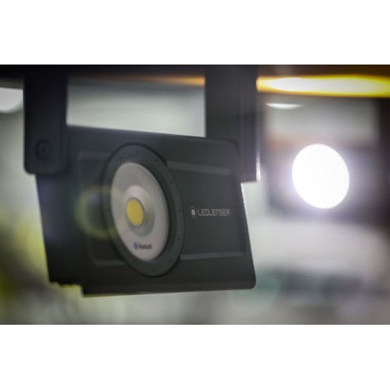 A close-up of a LEDLenser portable light, hanging from a surface, with its bright LED illuminated. The background is blurred, showing another light source and an indistinct setting, highlighting the focused and powerful beam of the LEDLenser.