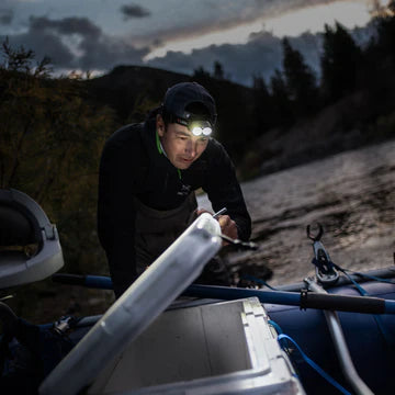 A person wearing a headlamp is leaning over an open container on a boat at dusk. The headlamp illuminates the container, allowing the individual to see its contents clearly. The person is dressed in outdoor gear, including a cap and a jacket, suggesting they are preparing for an outdoor activity such as fishing or camping. The background features a riverbank with trees and a cloudy sky, indicating an evening setting.