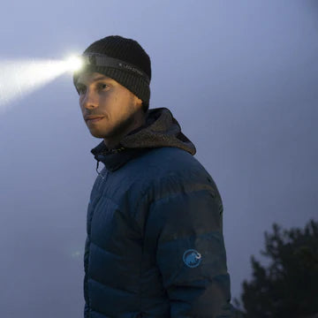A person wearing a blue jacket and a black beanie is using a headlamp that projects a bright beam of light. The setting appears to be outdoors in low-light conditions, with a misty or foggy background and trees faintly visible in the distance. The headlamp illuminates the path ahead, showcasing its effectiveness in dark environments.