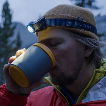 A person wearing a beanie and a LED Lenser headlamp is drinking from a mug. The headlamp is on, illuminating the area around them. The person is outdoors, dressed in warm clothing, suggesting a cold environment. Trees and mountains are faintly visible in the background, indicating a natural setting.
