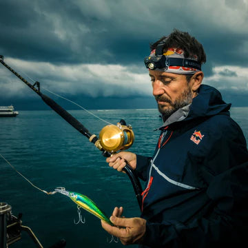 A man wearing a headlamp and a waterproof jacket is holding a fishing rod with a lure in his other hand. He is standing on a boat with a body of water and a cloudy sky in the background. The scene suggests that he is preparing for fishing, possibly in challenging weather conditions. The headlamp on his head is from LED Lenser, indicating he is equipped for low-light conditions.