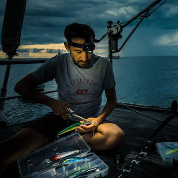 A man sitting on a boat's deck is wearing a headlamp and a t-shirt, focusing intently on preparing a fishing lure. The scene is set against a backdrop of a body of water under a cloudy sky, suggesting dusk or dawn. The man is using a pair of pliers and has a tackle box with various fishing lures next to him. The headlamp from LED Lenser provides illumination for his task in low-light conditions.