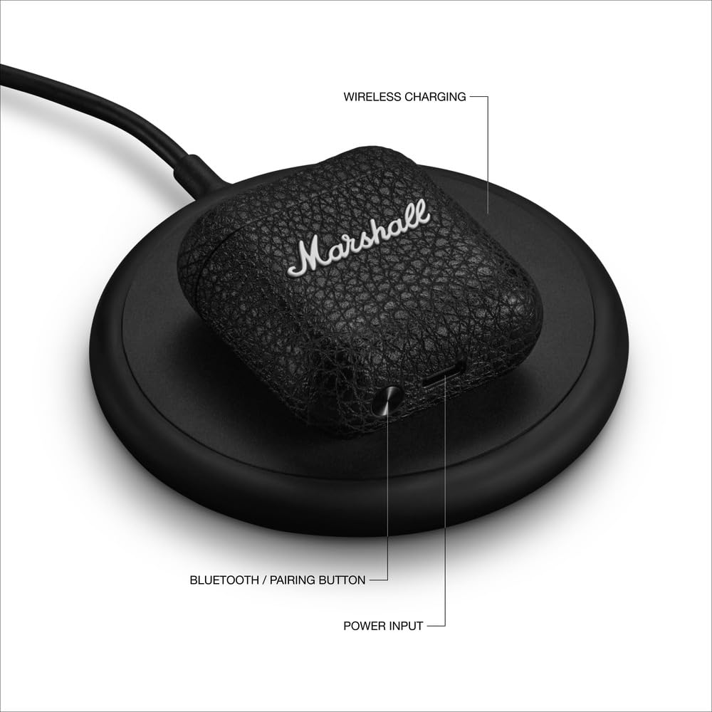 Marshall wireless earbuds charging case placed on a wireless charging pad. The case features a textured black finish with the Marshall logo on the front. Labeled components include the Bluetooth/pairing button and power input at the bottom of the case. The setup highlights the case's wireless charging capability for convenient power replenishment.