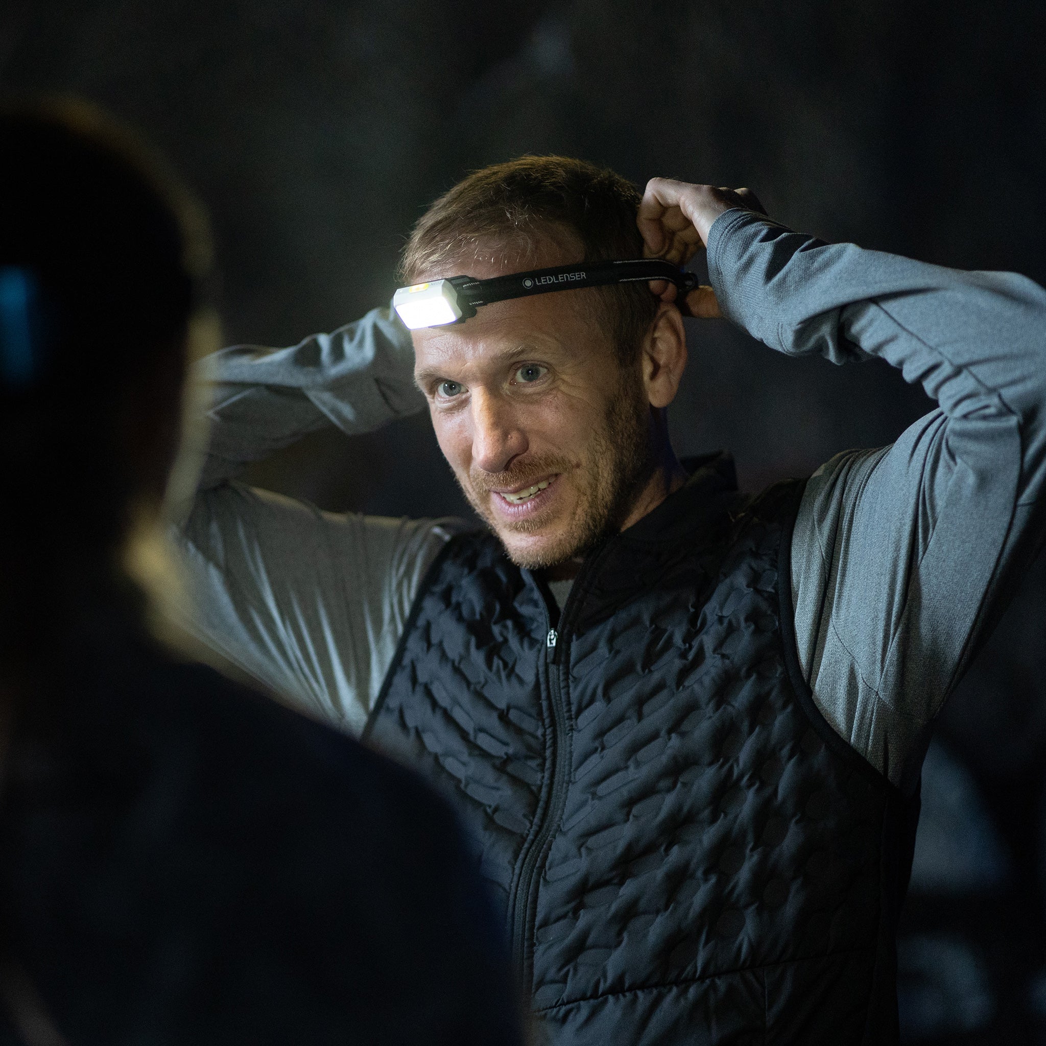 A man wearing a black quilted vest and a gray long-sleeve shirt adjusts a LED Lenser headlamp on his forehead. The headlamp is turned on, providing a bright light in the dark environment. Another person, slightly out of focus, is visible in the foreground. The setting suggests an outdoor or adventure activity at night.