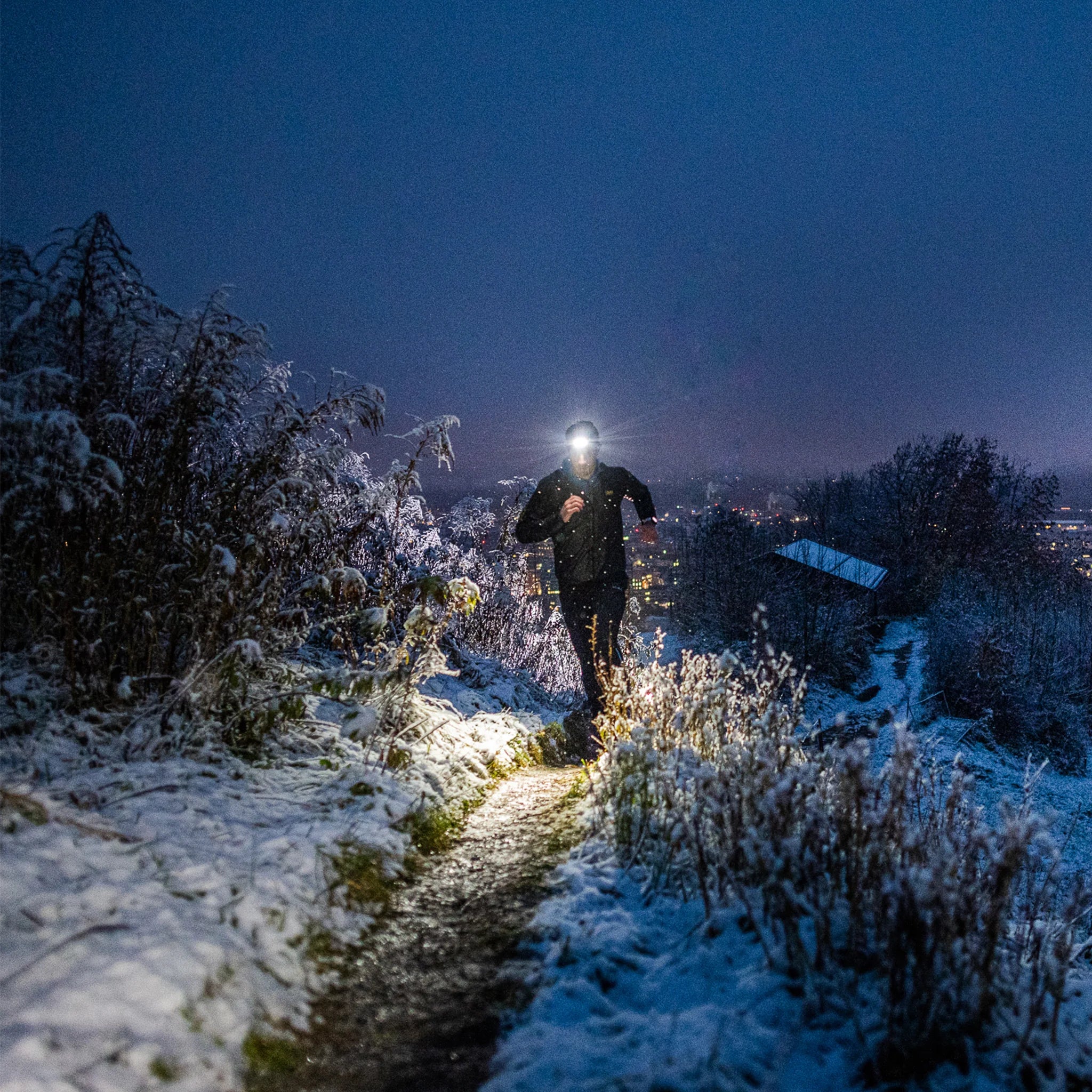 A person wearing a headlamp runs along a snow-covered path at dusk or dawn. The headlamp illuminates the trail ahead, while the background shows a town or city with lights twinkling in the distance. The scene captures the contrast between the dark sky and the brightly lit path, suggesting an outdoor adventure or exercise in cold weather.