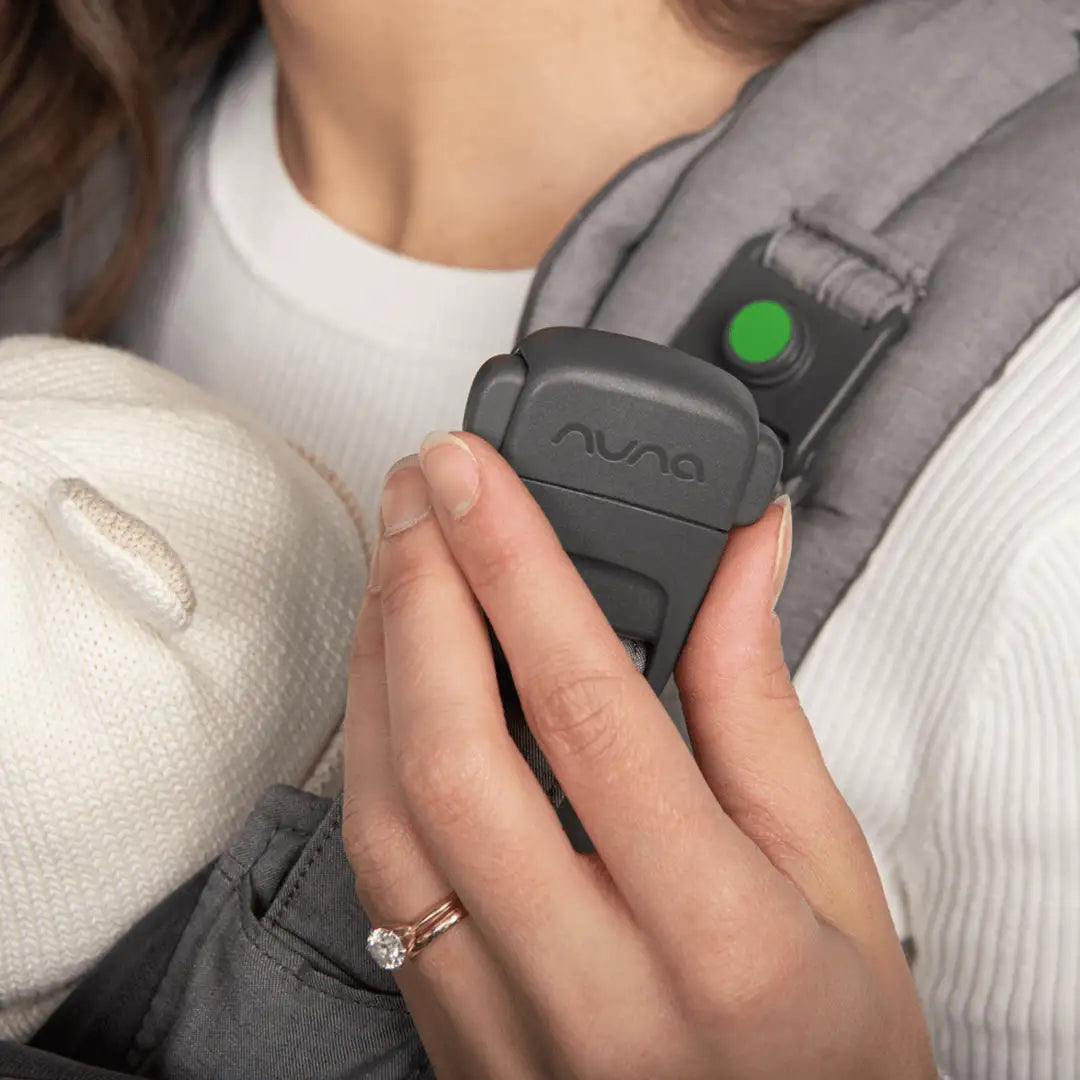 This image shows a close-up of a woman’s hand holding a black buckle with the brand name "Nuna" on it, part of a gray baby carrier strap. The carrier is worn over a white ribbed top. A green indicator is visible on the shoulder strap, likely showing that the buckle is securely fastened. A baby in a cream knit hat is partially visible, cradled close to the woman’s chest. The woman is wearing a diamond ring on her left hand.