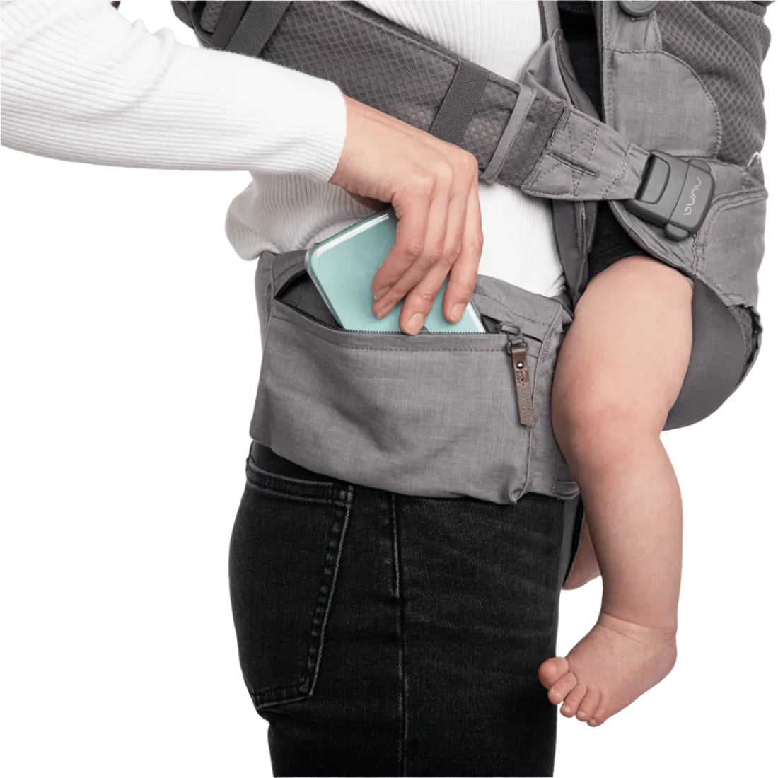This image shows a close-up view of a woman wearing a gray baby carrier. She is placing a mint green smartphone into a built-in zippered pocket located on the waistband of the carrier. A baby’s bare leg and foot are visible, showing that the child is securely seated in the carrier. The woman is dressed in a white top and black pants.