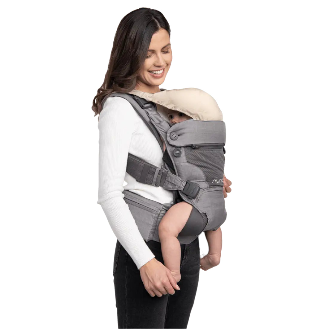This image shows a woman smiling while carrying a baby in a gray Nuna baby carrier. The baby is nestled comfortably facing the woman and partially covered by a light beige sunshade attached to the carrier. The woman is dressed in a white ribbed long-sleeve top and black pants. The carrier is securely fastened around her shoulders and waist with padded straps and buckles for support.