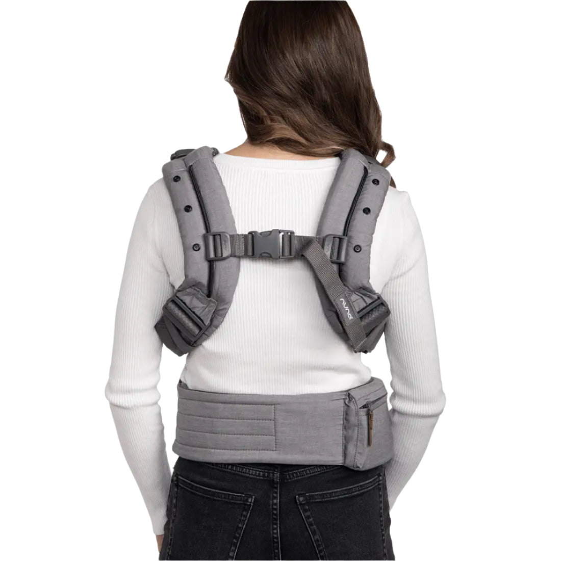 This image shows the back view of a woman wearing a gray Nuna baby carrier harness. The carrier has padded shoulder straps secured with a chest clip and adjustable side straps. A zippered pouch is attached to the waistband, which wraps snugly around her waist. She is dressed in a white ribbed long-sleeve top and black jeans.