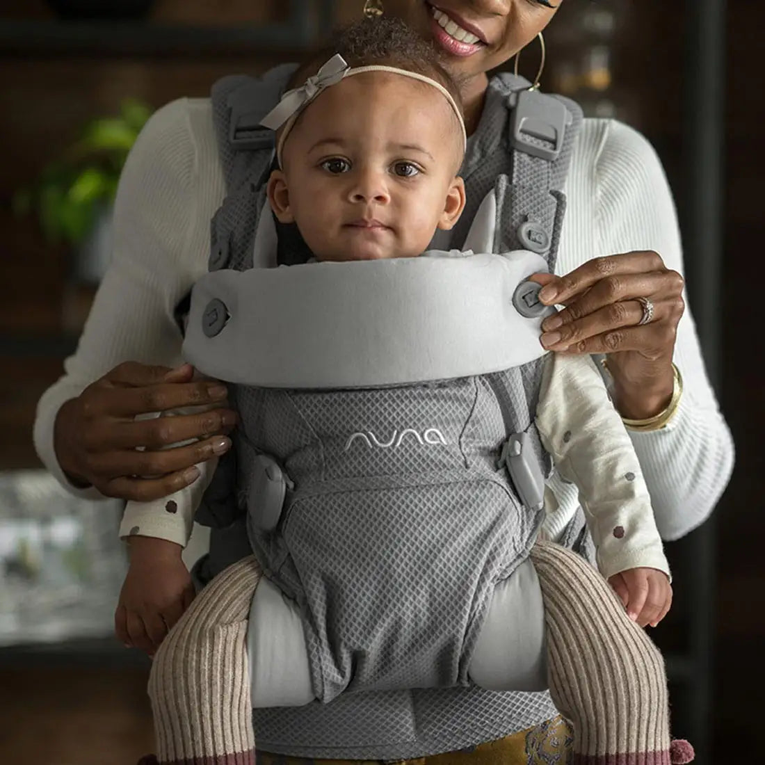 This image shows a mother holding her baby in a gray Nuna baby carrier. The baby is facing forward, wearing a white long-sleeve shirt and light-colored tights. The carrier has a soft gray headrest with buttons and is securely strapped around the mother’s body.