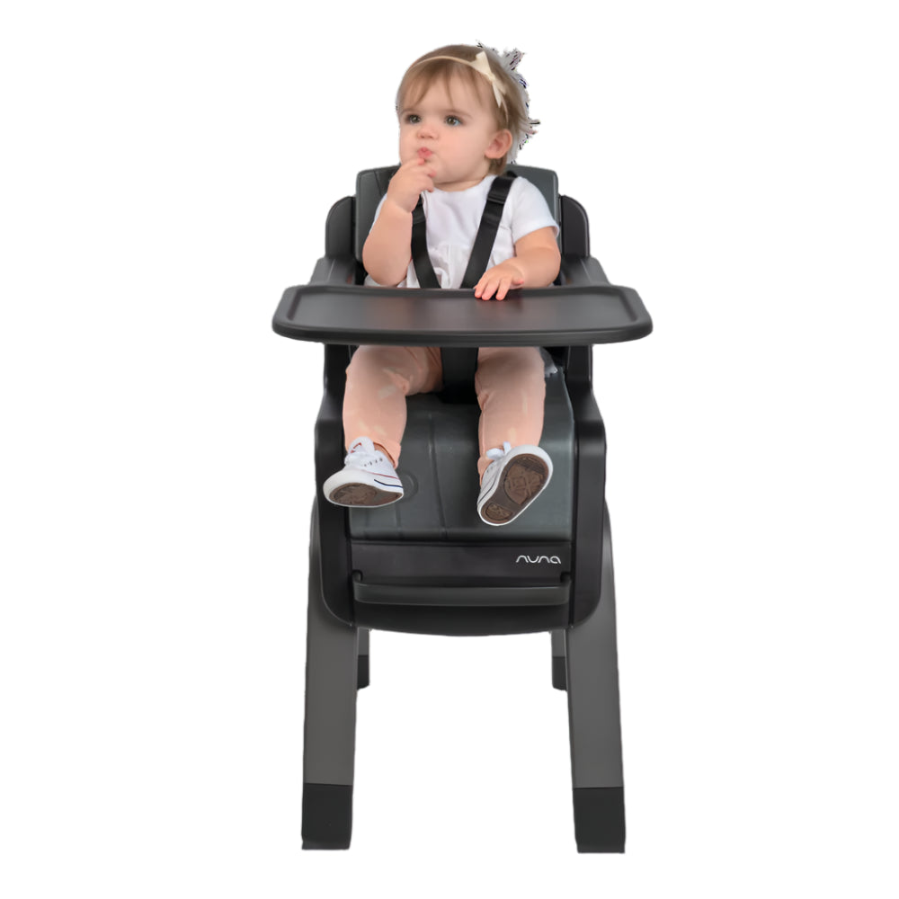 Toddler sitting in a black Nuna high chair with safety harness and tray, wearing a white shirt and pink pants, appearing curious while eating.