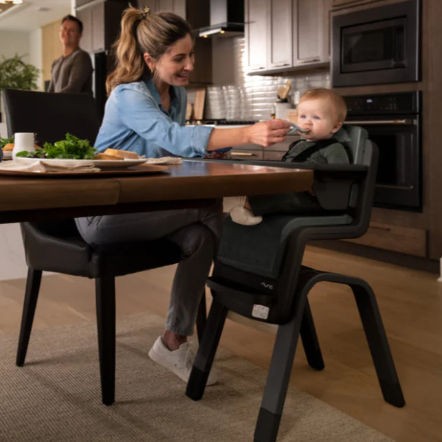 Mother feeding her baby seated in a Nuna high chair at a dining table in a modern kitchen, with the baby secured by a harness and smiling during mealtime.