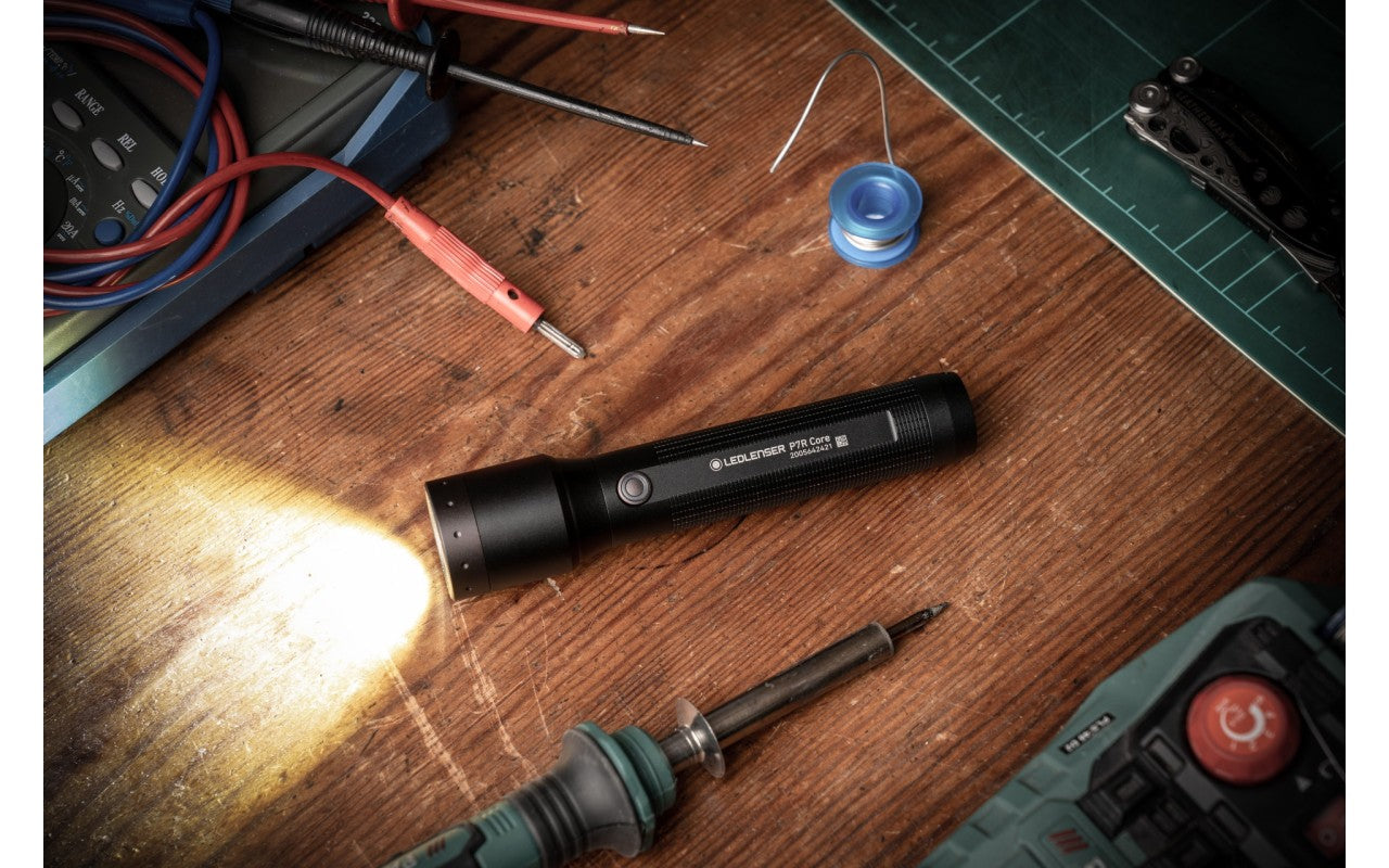A black LED flashlight labeled "LEDLENSER P7R Core" lying on a wooden workbench. The flashlight is on, casting a bright light to the left. Surrounding the flashlight are various tools, including a soldering iron, a roll of solder wire, a multimeter with attached probes, and a utility knife. The workbench also has a green cutting mat in the upper right corner, indicating a workspace for electronic or mechanical tasks.