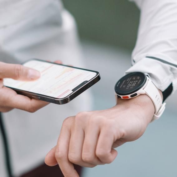 A close-up of a person wearing a Polar Pacer smartwatch with a white strap on their left wrist, holding a smartphone in their right hand. The smartwatch screen is visible, displaying information, and the person is interacting with the smartphone. The person is dressed in a light-colored jacket, and the background is blurred, emphasizing the watch and the phone.