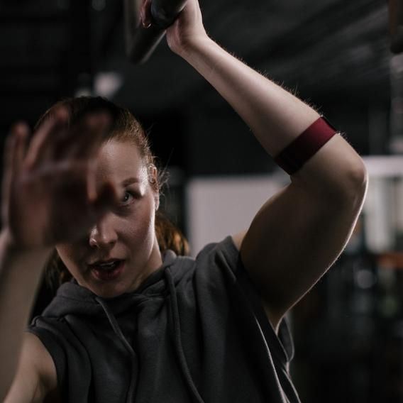 A person working out in a gym, wearing a Polar optical heart rate sensor on their upper arm, secured with a red adjustable strap. The individual is dressed in a sleeveless hoodie and is performing an exercise with their arms raised, emphasizing the sensor's placement and use during physical activity. The gym environment is visible in the background, highlighting the fitness context.