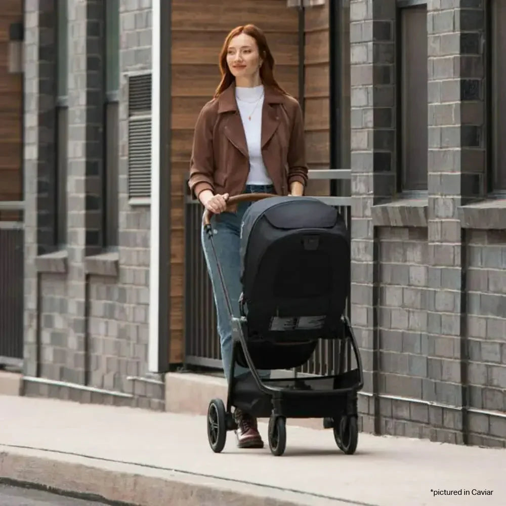 A woman is pushing a modern black stroller with a sleek frame and large canopy while walking along a city sidewalk.
