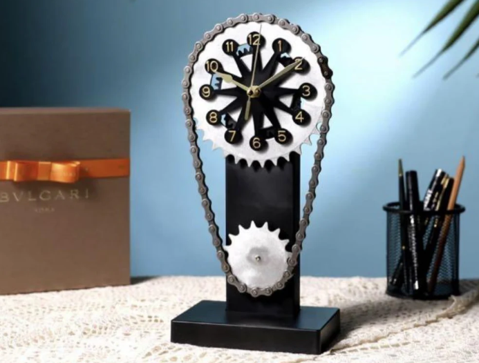 Gear-inspired clock with a chain-driven design, displayed on a lace-covered table against a soft blue background. The setup includes a gift box with an orange ribbon and a pen holder, creating a stylish and elegant scene.