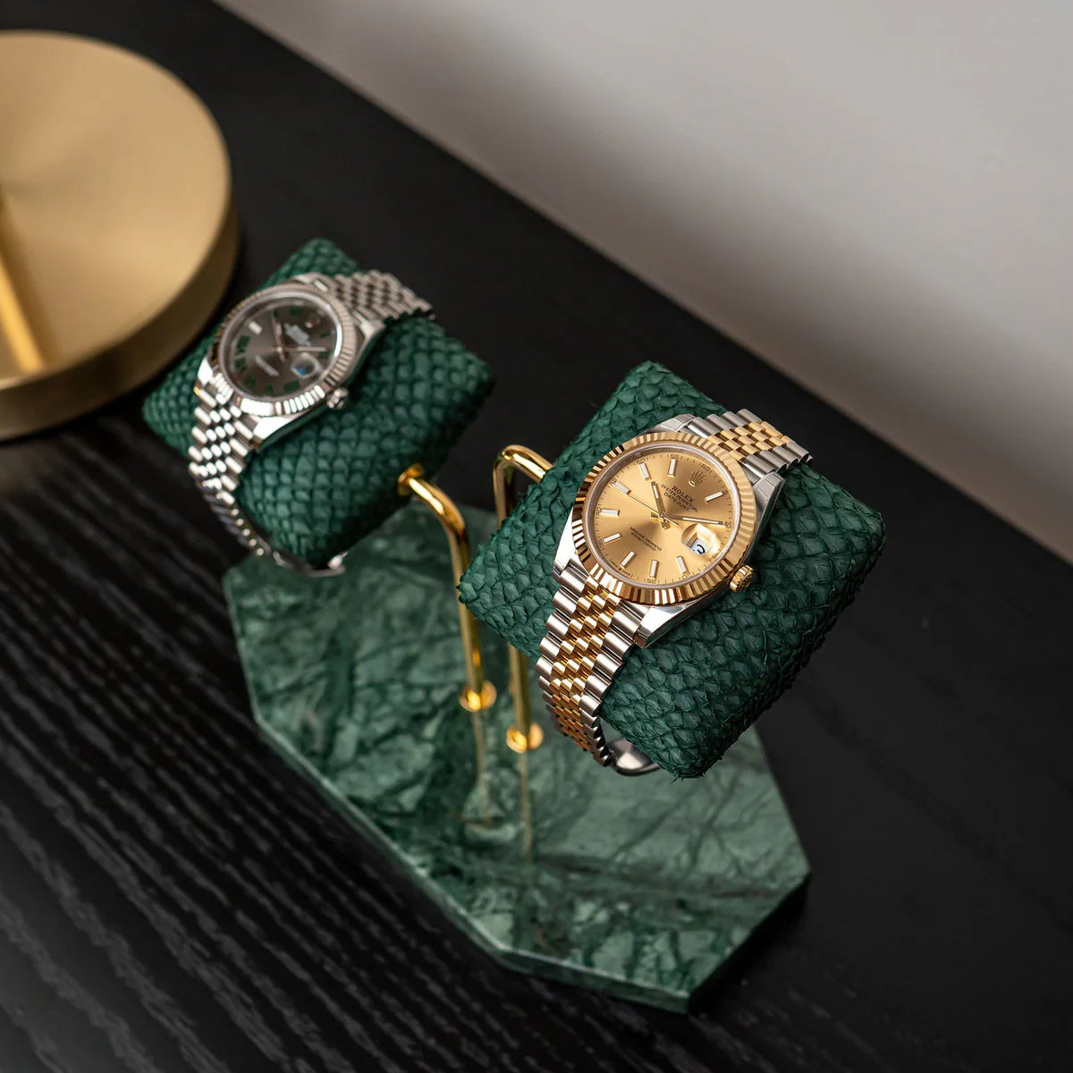 Two luxurious wristwatches are displayed on green textured cushions, mounted on a double-arm stand with a green marble base. The stand features elegant gold supports, adding to the sophisticated design. One watch has a silver metal band with a silver dial, while the other has a two-tone metal band (gold and silver) with a gold dial. The display is set on a dark wooden surface, with a gold lamp partially visible in the background, creating a stylish and refined presentation.