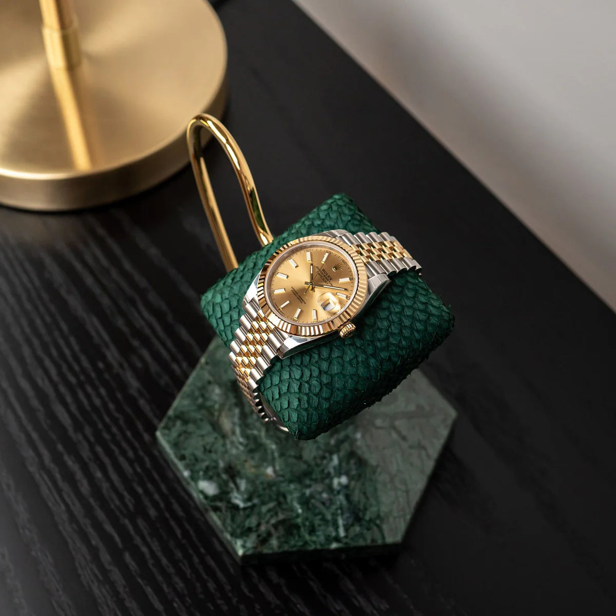A luxurious gold wristwatch with a silver and gold metal band is displayed on a green textured cushion. The cushion is attached to a gold stand with a marble base, placed on a dark wooden surface. The watch face is gold, complementing the elegant display setup. In the background, part of a gold lamp is visible, adding to the sophisticated ambiance.