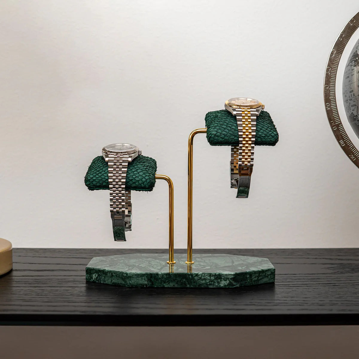 Two luxurious wristwatches are displayed on green textured cushions, mounted on a double-arm stand with a green marble base. The stand features elegant gold supports, adding to the sophisticated design. One watch has a silver metal band with a silver dial, while the other has a two-tone metal band (gold and silver) with a gold dial. The display is set on a dark wooden surface against a plain white wall, creating a stylish and refined presentation.