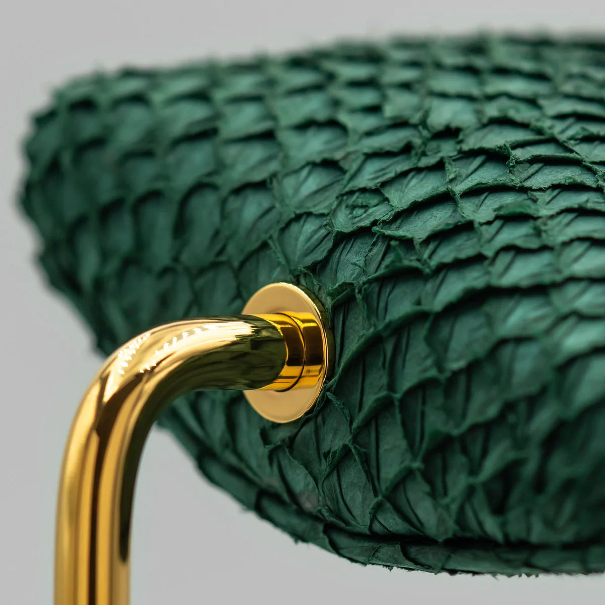 A close-up view of a green textured cushion attached to a shiny gold curved metal support. The cushion has a scaly, reptile-like texture, and the metal support is secured with a polished gold screw. The image highlights the luxurious materials and the detailed craftsmanship of the display stand.