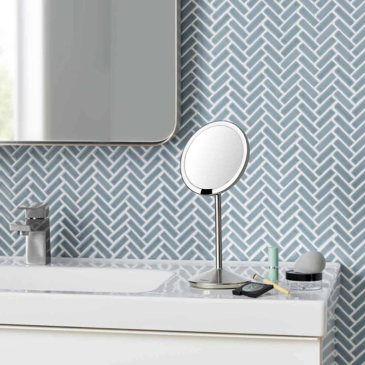 A modern bathroom countertop featuring a sleek, circular vanity mirror with a brushed stainless steel stand. The countertop is made of marble and has a few items placed on it, including a toothbrush in a holder, a small container, and a smartphone. The wall behind the countertop is adorned with blue and white herringbone-patterned tiles, adding a stylish and contemporary touch to the bathroom. A partial view of a wall-mounted mirror is visible above the countertop, reflecting natural light, contributing to