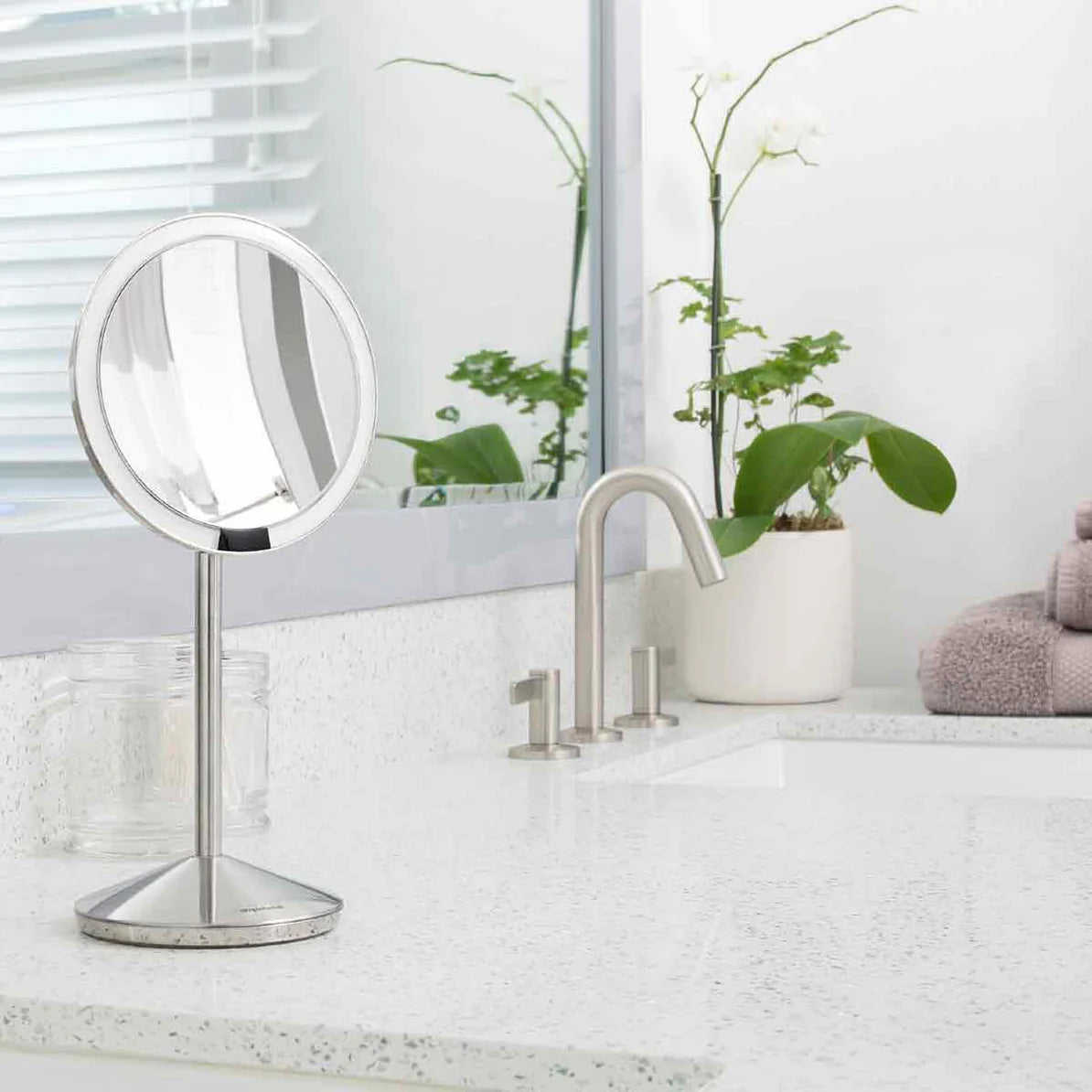A modern bathroom countertop with a clean, white, speckled surface. On the countertop is a sleek, circular vanity mirror with a brushed stainless steel stand. The mirror is positioned near a stylish faucet with a minimalist design. In the background, there is a potted plant with green leaves, adding a touch of nature to the space. A set of neatly folded towels and a glass jar are also visible on the countertop, contributing to the organized and serene ambiance of the bathroom. The overall aesthetic is fres