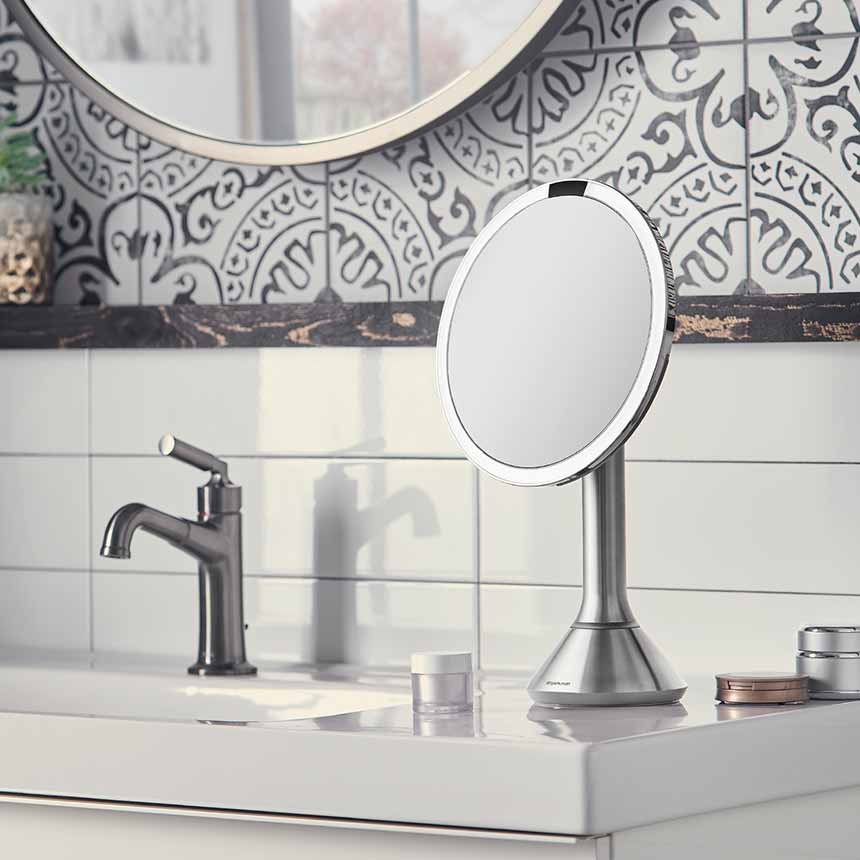 This image features a **Simplehuman makeup mirror** placed on a bathroom vanity. The mirror has a sleek, stainless steel finish, which complements the modern and sophisticated decor of the bathroom. The LED light ring around the mirror provides bright, even illumination, perfect for makeup application or grooming tasks. The mirror is set against a backdrop of decorative tiles with a stylish, intricate pattern, adding to the overall aesthetic of the space. The vanity also includes other elements like a fauc