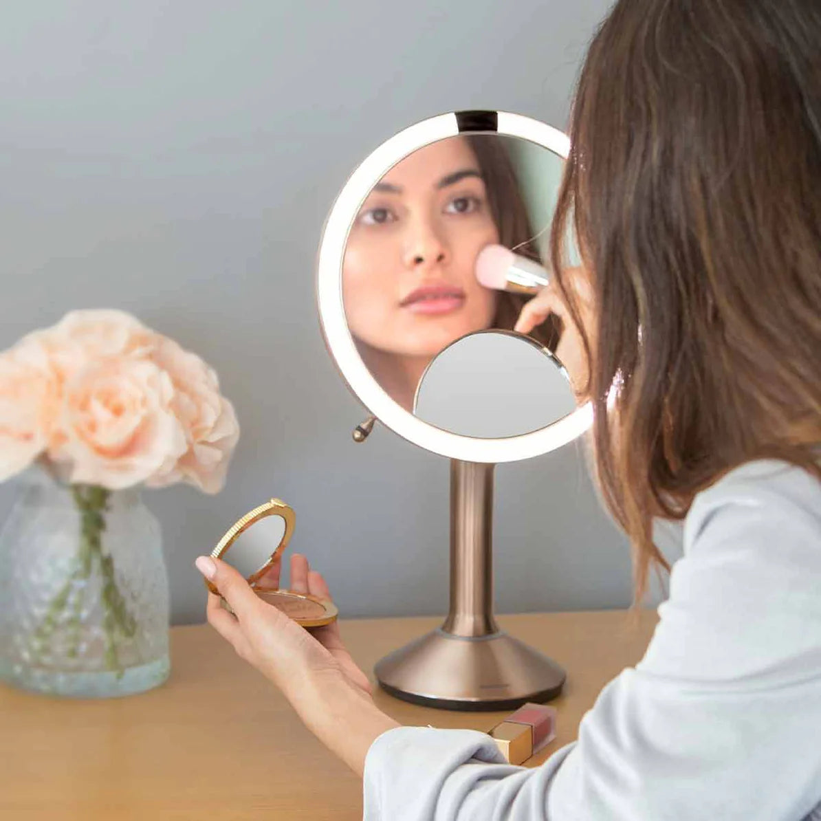 This image shows a woman using the Simplehuman makeup mirror as she applies makeup. The mirror features a bright LED light ring, providing clear, even illumination that enhances visibility. The reflection in the mirror highlights the mirror's ability to deliver true-to-life color and detail, which is essential for precise makeup application. The mirror is placed on a vanity, where the woman is using it to blend makeup with a brush while holding a compact in her other hand. The design of the mirror stand is