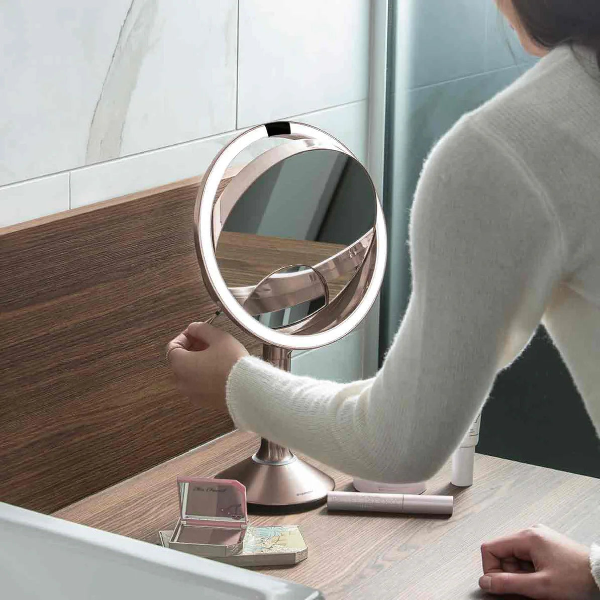 This image shows a person adjusting a Simplehuman makeup mirror on a wooden vanity or countertop. The mirror features a bright LED light ring and a sleek, rose gold metallic finish, which adds a touch of sophistication to the setup. The user appears to be adjusting the tilt or rotation of the mirror, highlighting its flexibility and user-friendly design. On the vanity, there are various beauty products, including makeup items such as a compact, lipstick, and possibly a skincare product, suggesting that thi