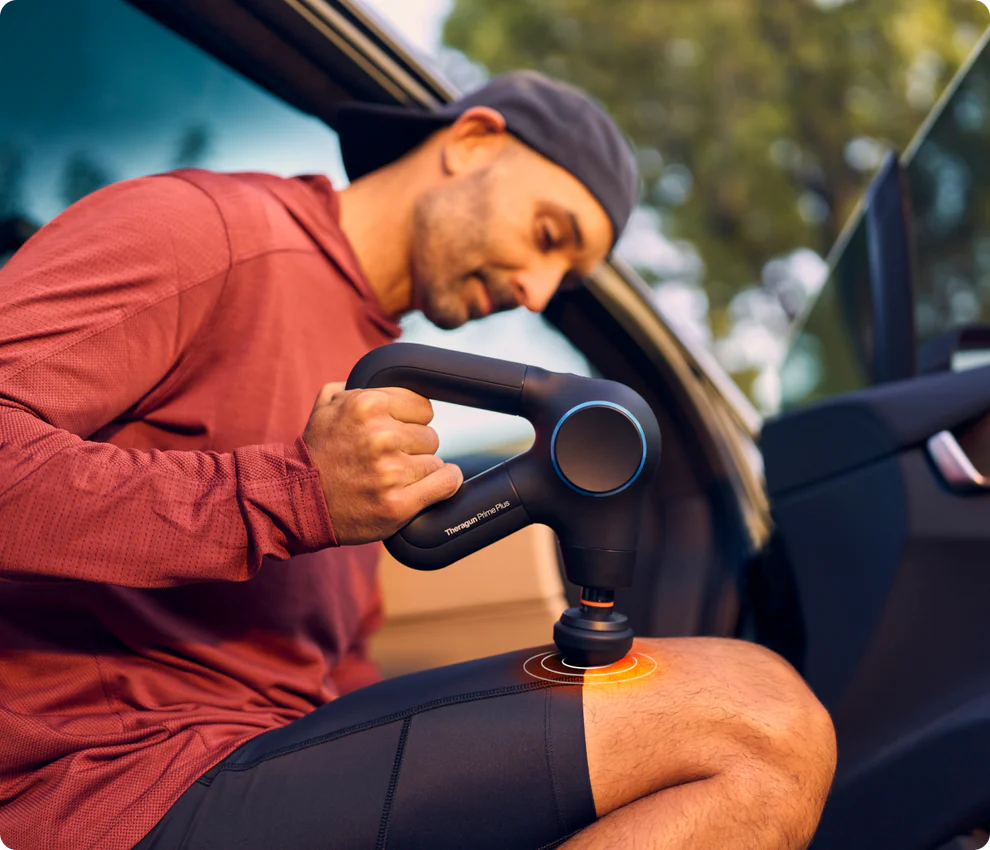 A man in a red hoodie and black cap sits in the open doorway of a car, using a black Theragun Pro Plus massage gun on his thigh. The device emits a glowing effect at the contact point, highlighting muscle relief. The background features blurred greenery, suggesting an outdoor or post-exercise recovery setting.