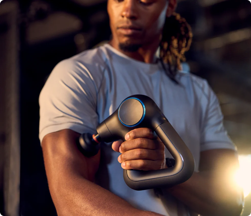 A fit man in a gray athletic shirt uses a black Theragun massage gun on his upper arm. The massage gun has a triangular handle and a round attachment, emitting a subtle glow, indicating use. The background is dimly lit, suggesting a gym or workout setting.