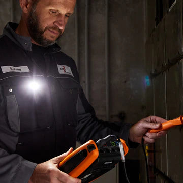 A man wearing a work uniform with a name tag reading "Steve" is using an LED clip light attached to his chest pocket to illuminate his work area. He is holding an orange and black electronic device in one hand and an orange tool in the other, indicating he is performing a technical task or inspection. The LED light provides bright, focused illumination in the dimly lit environment.